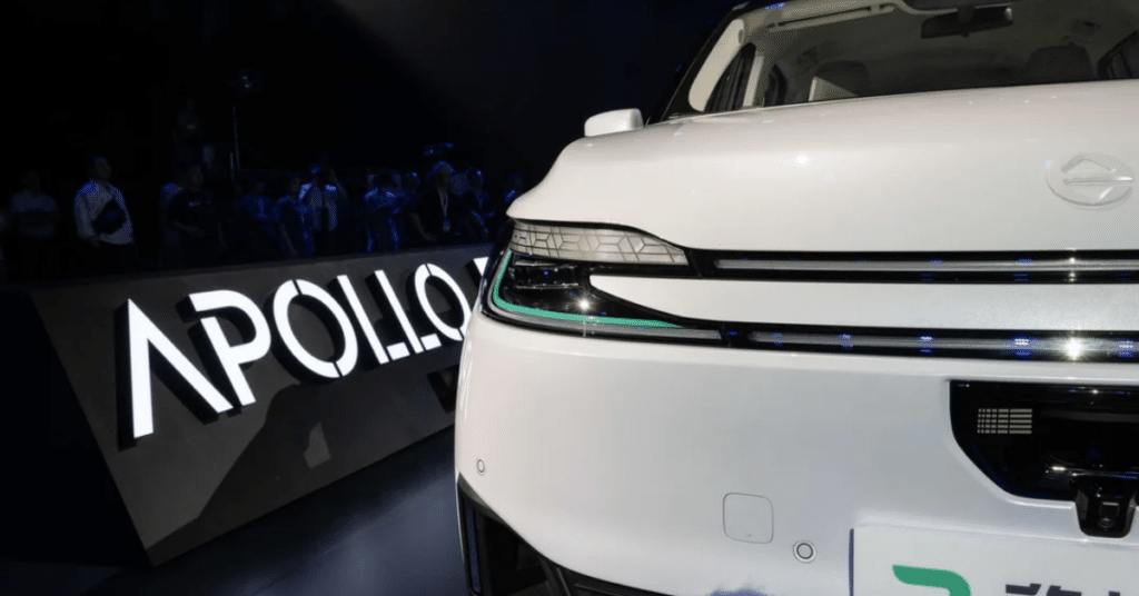 Close-up of a white Apollo autonomous vehicle on display, with sleek LED headlights illuminated and the “APOLLO” logo glowing in the background at a launch event.