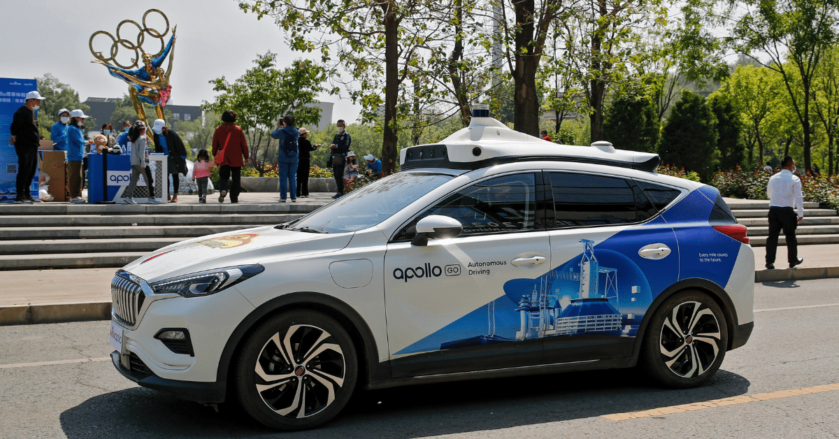 An autonomous SUV branded “Apollo Go Autonomous Driving” drives past spectators near an Olympic rings sculpture in a park-like urban setting.