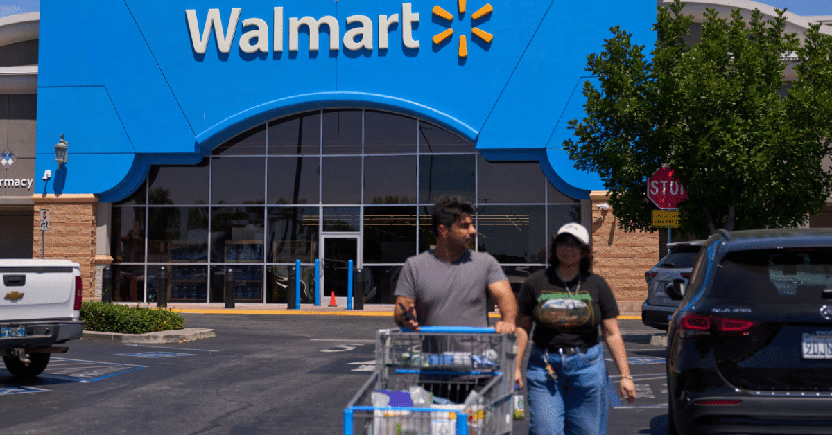 Shoppers push a cart outside a Walmart store entrance with the Walmart logo prominently displayed on a bright blue facade.