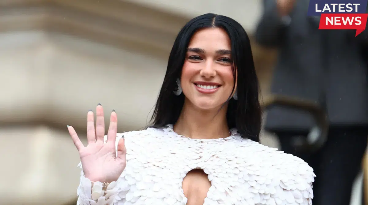 A smiling woman with long black hair waves to the camera while wearing a white textured outfit at a public appearance event.A smiling woman with long black hair waves to the camera while wearing a white textured outfit at a public appearance event.